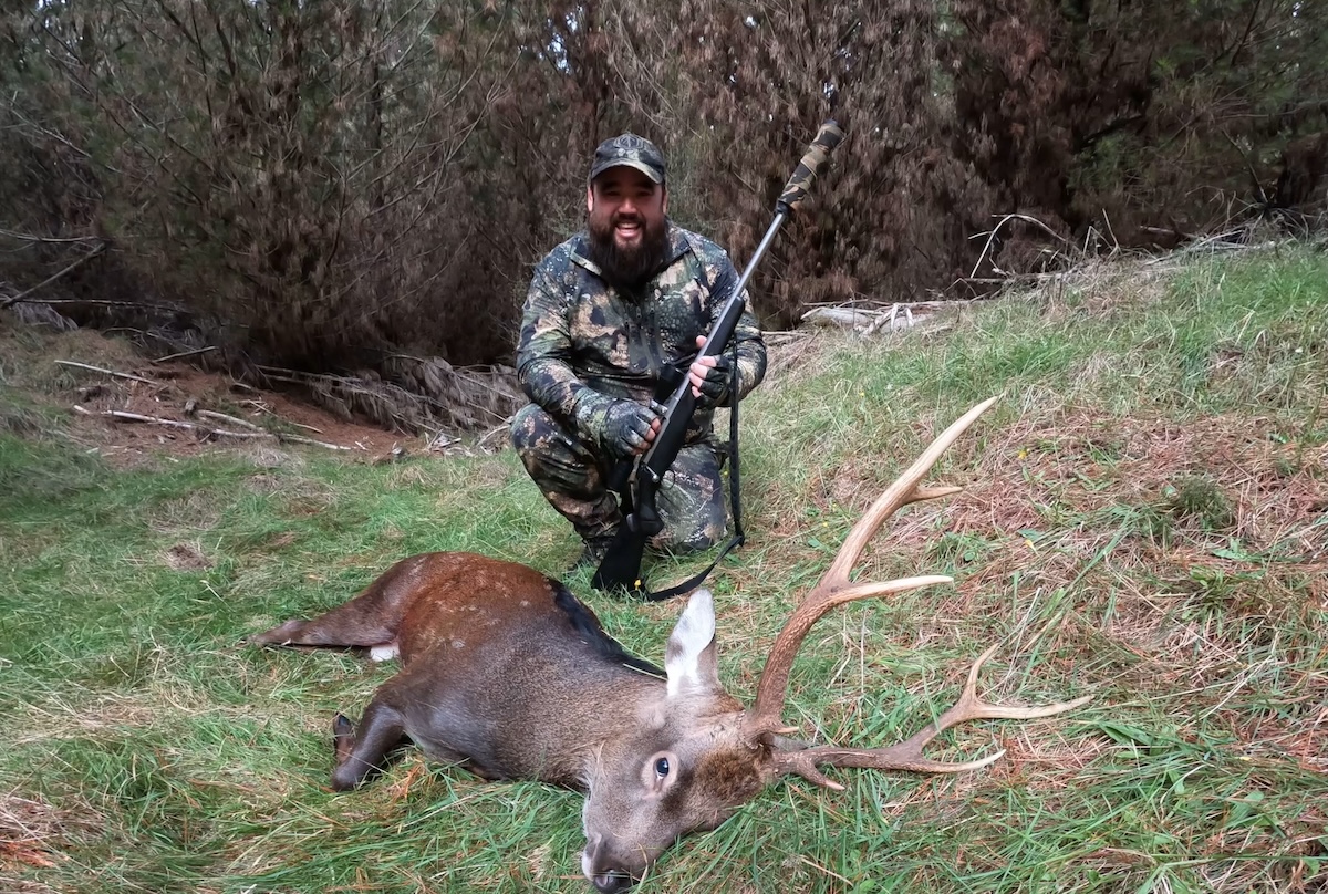 A bearded man wearing full camouflage hunting gear, including a jacket, pants, gloves, and a cap, is kneeling on a grassy slope in a forested area. He is holding a bolt-action rifle with a suppressor and has a broad smile on his face. In front of him lies a recently harvested stag with large antlers, resting on the grass. The background consists of a dense tree line with dried branches and foliage.
