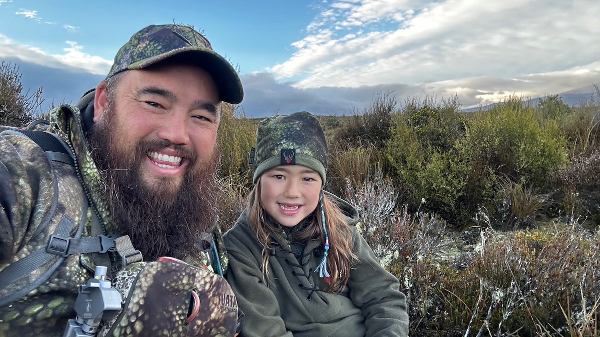 A bearded man and a young girl are sitting outdoors in a rugged, natural landscape with low-lying shrubs and an overcast sky. They are both smiling at the camera, dressed in camouflage hunting gear, including jackets, hats, and backpacks. The man has a thick beard and is wearing a camo cap, while the girl has long hair with colorful braided strands and is wearing a matching beanie with a deer logo. The gear has a 'Tuatara Camo' pattern. The background consists of an open wilderness area with rolling terrain and a mix of green and brown vegetation.