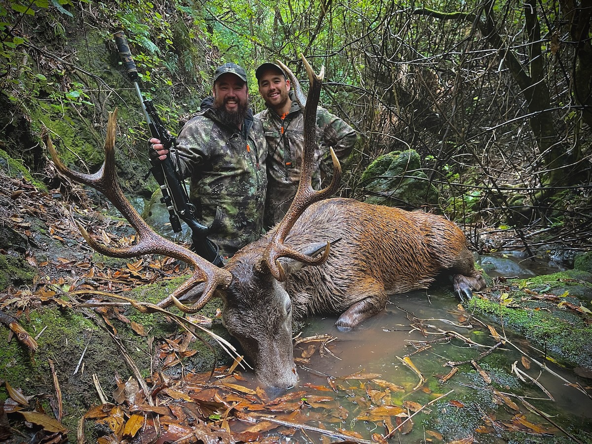 Two bearded men dressed in camouflage hunting gear are posing in a dense forest next to a large red stag lying partially submerged in a shallow creek. The man on the left is holding a bolt-action rifle with a suppressor and has a broad smile, while the man on the right stands slightly behind him, also smiling. The surrounding environment is lush with moss-covered rocks, fallen leaves, and tangled branches overhead, creating a rugged, natural setting.