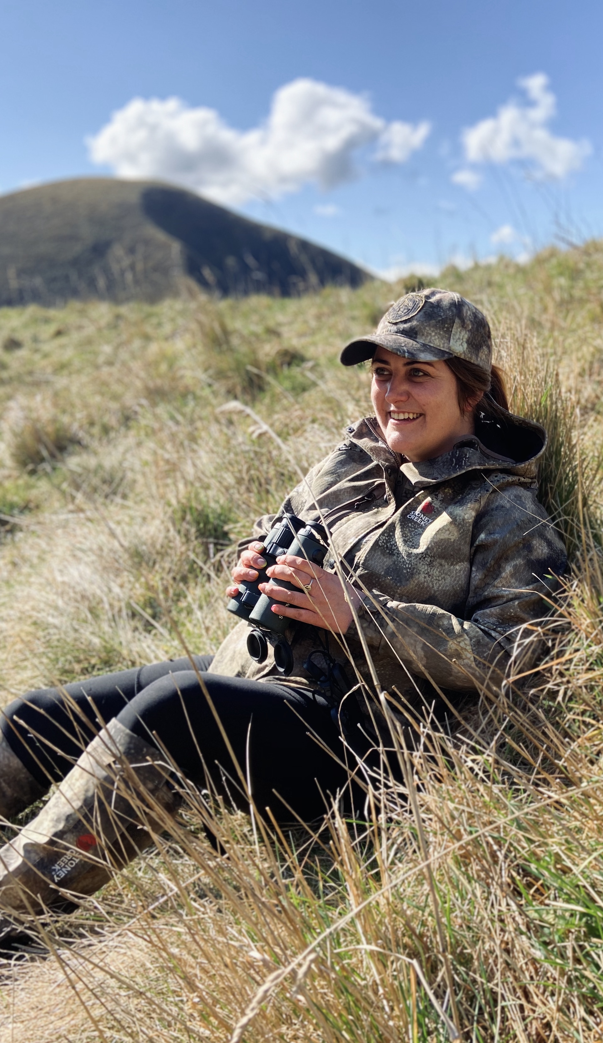 A hunter dressed in full camouflage gear, including a jacket, pants, and a cap, is kneeling next to a large red stag lying on the ground. The hunter is holding a bolt-action rifle and smiling at the camera. The background consists of a rugged outdoor landscape with dry grass, shrubs, and rolling hills, under an overcast sky.